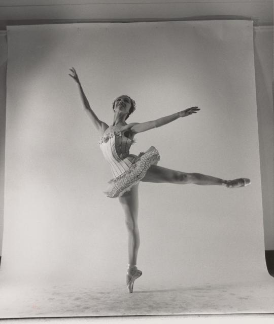 A black and white promotional photo of Maria Tallchief on pointe with one leg and both arms up in the air. The edges of the backdrop can be seen.