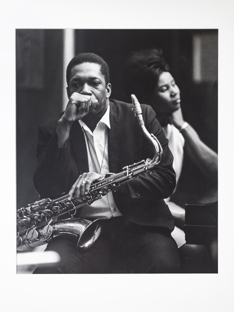 Black and white photograph showing a seated Black man holding a saxophone, with a Black woman sitting behind him at a piano. 
