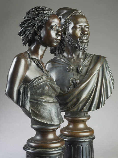 A pair of sculpted busts: they show the head and shoulders of a Black woman and man, each supported on a circular pedestal or short pillar. Both the busts and the pedestals are made of dark brownish colored bronze. 