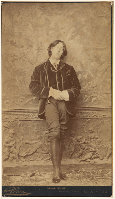 Portrait of Oscar Wilde in a velvet vest and jacket and tall boots leaning against an ornate wall by Napoleon Sarony.