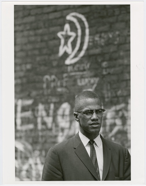 A photograph of Minister Malcolm X in Harlem, New York, with a graffiti star and crescent moon on a brick wall behind him. 