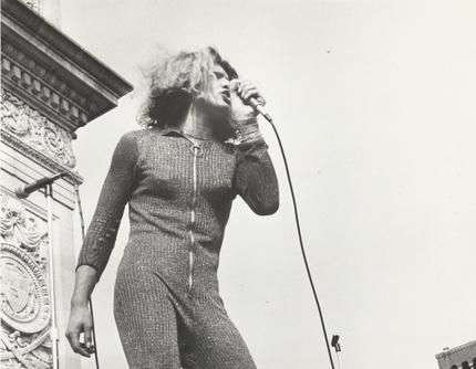 Photograph of Sylvia Rivera holding a microphone, speaking in front of Washington Square arch