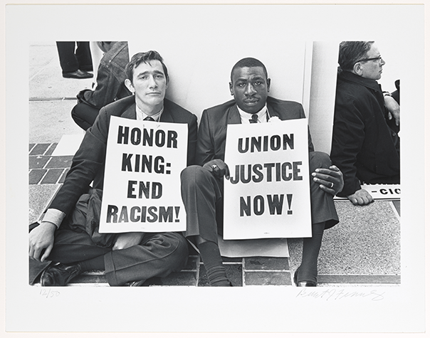  [Alternative text] Black-and-white photograph of two men seated on the ground, one holding a placard reading “Honor King: End Racism!” and the other “Union Justice Now!”
