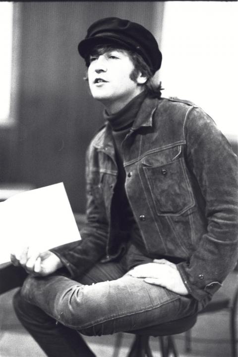 In a 1965 black and white photograph John Lennon sits crosslegged on a stool in a reccording studio. 