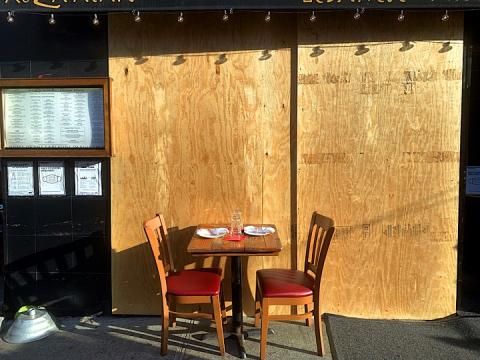 A dining table and two chairs with place settings on the sidewalk in front of boarded up windows