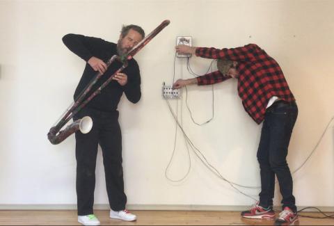 Two men playing homemade instruments in front of a white wall.