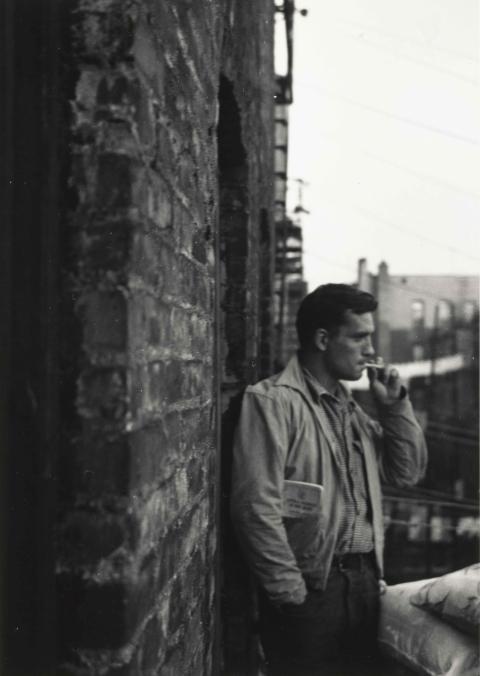 Black and white photograph of Jack Kerouac standing in profile against a building, smoking a cigarette.