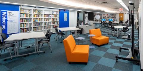 Interior of Best Buy Teen Tech Center at Grand Concourse Library, featuring blue checkered carpet, orange lounge chairs in front of a monitor screen, shelves of books, and work tables.